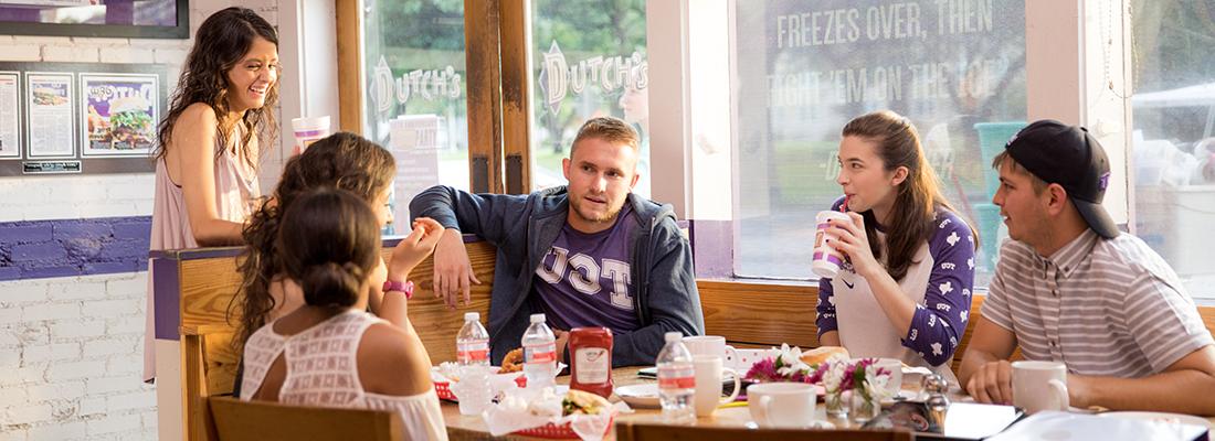 Off campus, a group of six TCU students laugh and eat hamburgers together at a large table at Dutch's burgers.